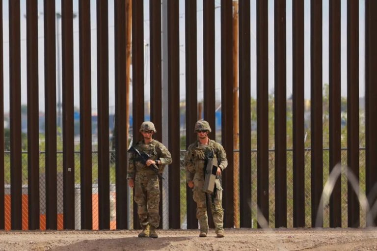 Two military guards in full uniform holding anti-drone equipment in front of a tall border wall fence.