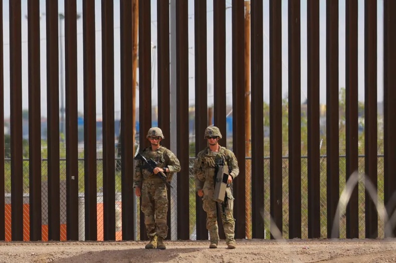 Two military guards in full uniform holding anti-drone equipment in front of a tall border wall fence.