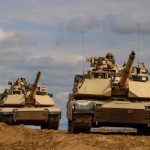 Two military tanks driving on a dirt terrain under a cloudy sky.
