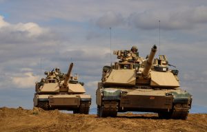 Two military tanks driving on a dirt terrain under a cloudy sky.
