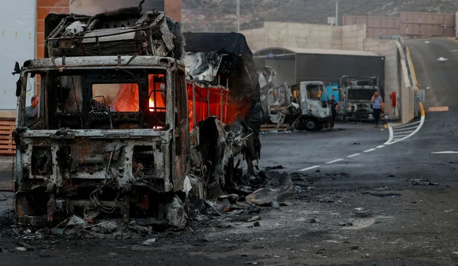 A heavily burned and destroyed truck on a road with damaged vehicles and emergency personnel nearby.