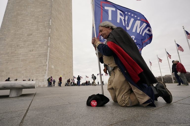A supporter dressed in colonial attire kneeling near the Washington Monument holding a Trump flag.