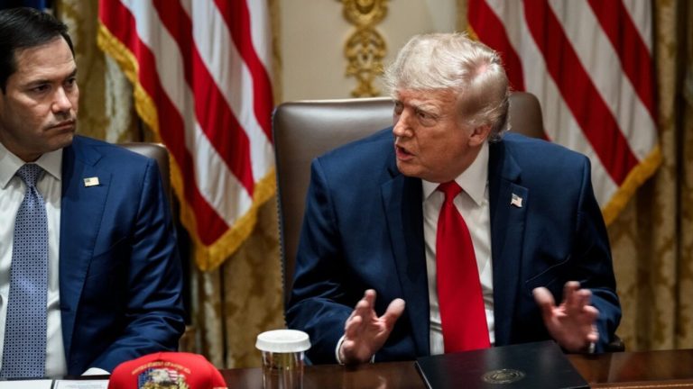 Donald Trump speaking while seated at a conference table next to Marco Rubio in front of U.S. flags.