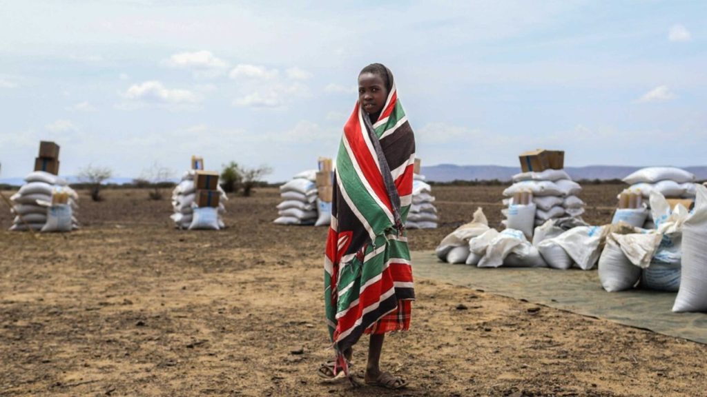 A child wrapped in a patterned blanket standing near aid supplies in an arid landscape.