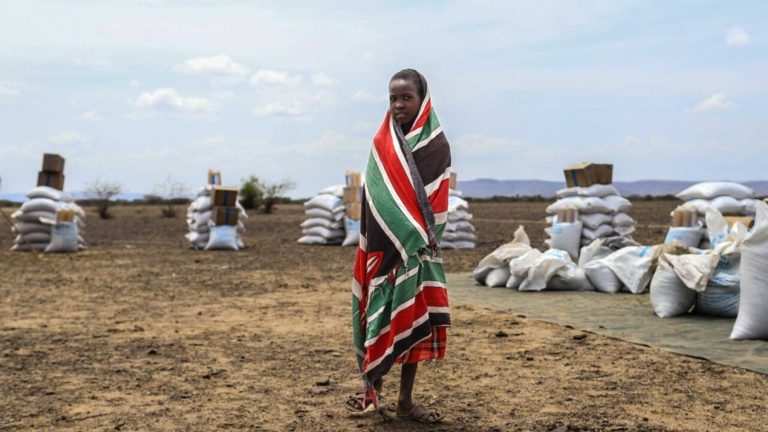A child wrapped in a patterned blanket standing near aid supplies in an arid landscape.