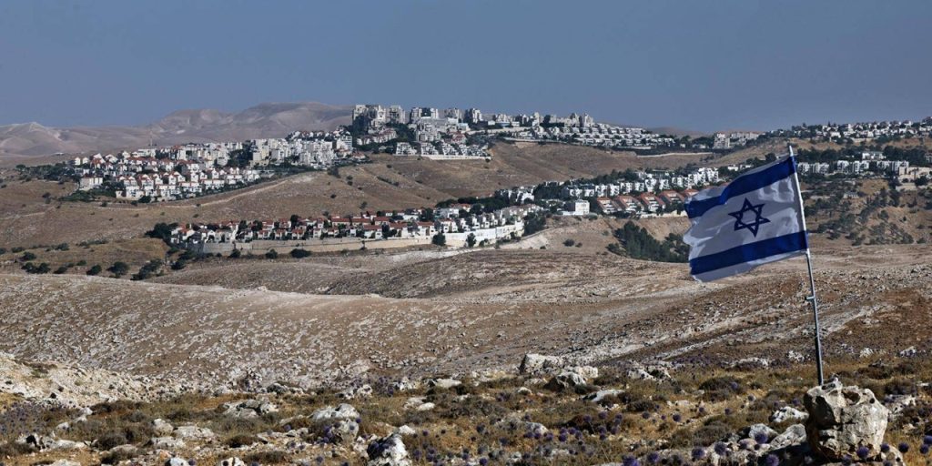 An Israeli flag waving on a rocky hilltop overlooking a residential settlement built across dry hills.