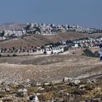 An Israeli flag waving on a rocky hilltop overlooking a residential settlement built across dry hills.