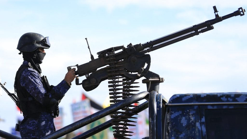 Armed fighter in combat gear standing behind a mounted heavy machine gun on a military vehicle.