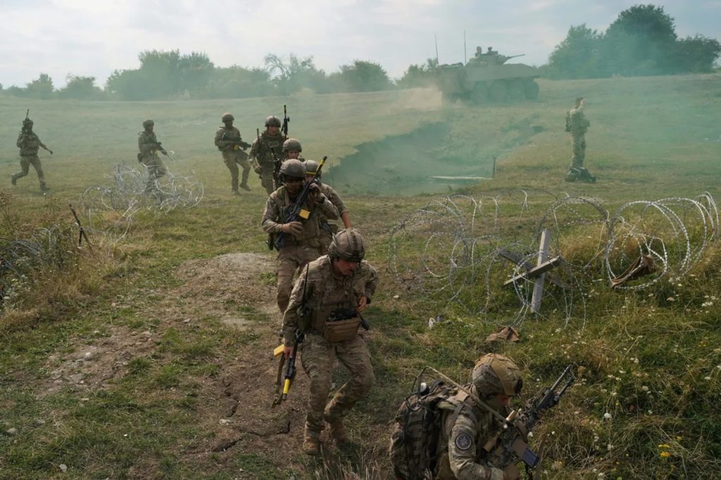 Military infantry soldiers maneuvering through field obstacles and barbed wire during a tactical exercise.