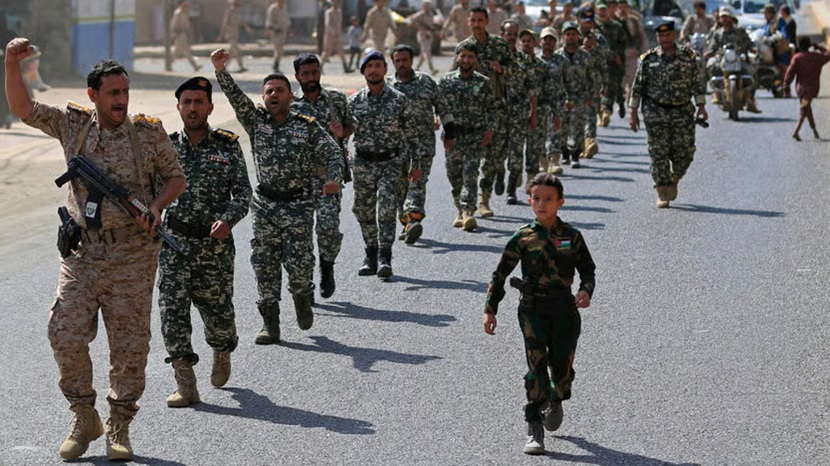Uniformed soldiers marching down a street alongside a young boy in military fatigue attire.