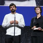 Palestinian and Israeli filmmakers speaking at a microphone while holding an award at a film festival stage.