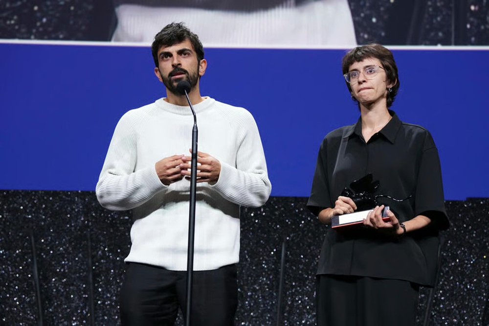Palestinian and Israeli filmmakers speaking at a microphone while holding an award at a film festival stage.