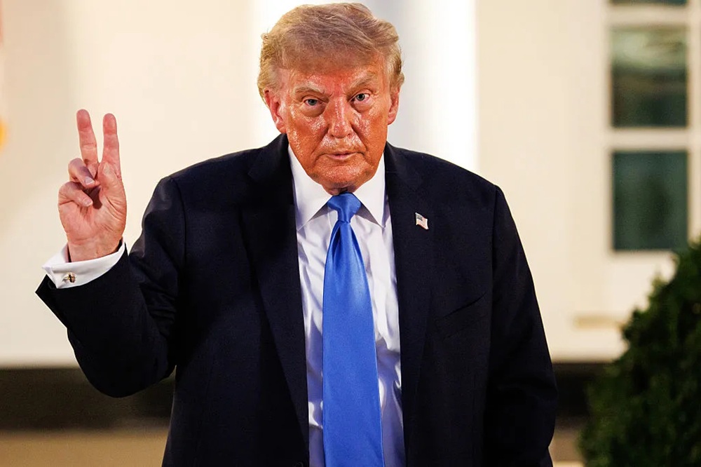 Former U.S. President Donald Trump making a peace sign gesture while wearing a suit and blue tie.