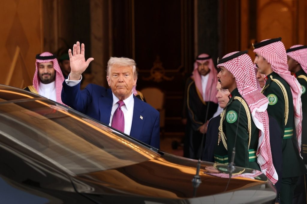 Donald Trump waving next to an official motorcade vehicle alongside Saudi military personnel.