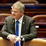 Israeli politician in a dark suit wearing glasses leaning against a leather seat in a parliament assembly room.