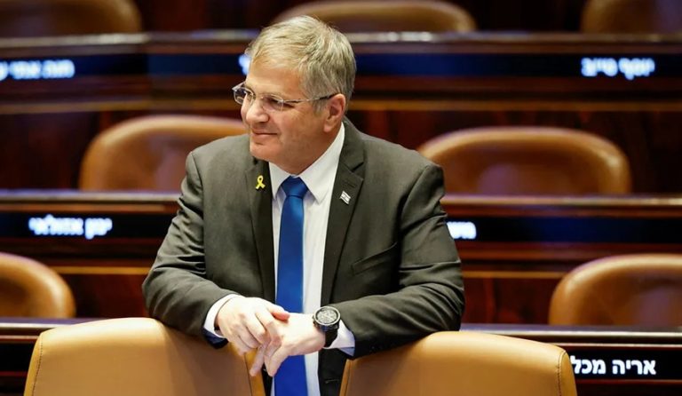 Israeli politician in a dark suit wearing glasses leaning against a leather seat in a parliament assembly room.