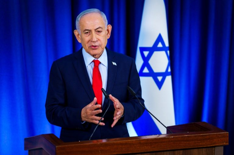 Israeli Prime Minister Benjamin Netanyahu gesturing with both hands while speaking at a wooden podium in front of the flag of Israel.
