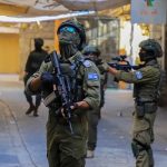 Armed military personnel in tactical gear patrolling a narrow urban street.