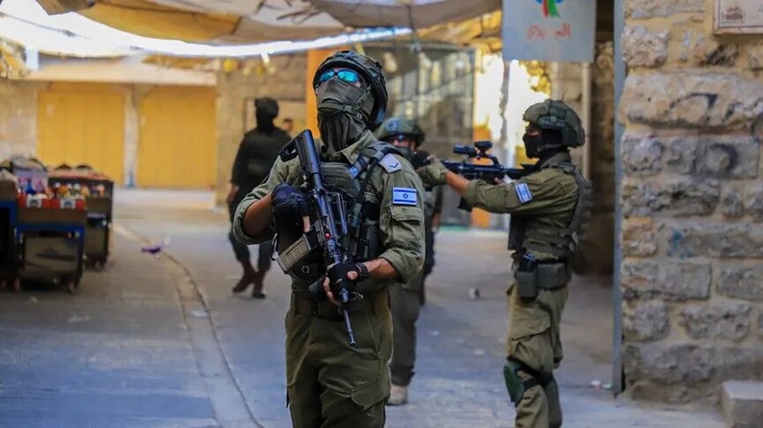 Armed military personnel in tactical gear patrolling a narrow urban street.