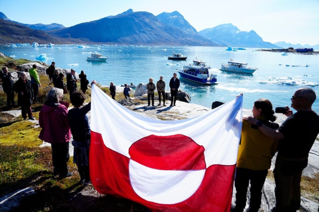 People holding a large Greenlandic flag overlooking an Arctic fjord with boats and icebergs.
