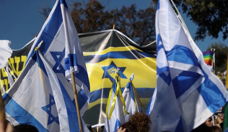 Demonstrators waving Israeli national flags and political banners at an outdoor political rally.