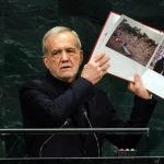 Iranian President Masoud Pezeshkian holding up an open photo book at the UN General Assembly podium.