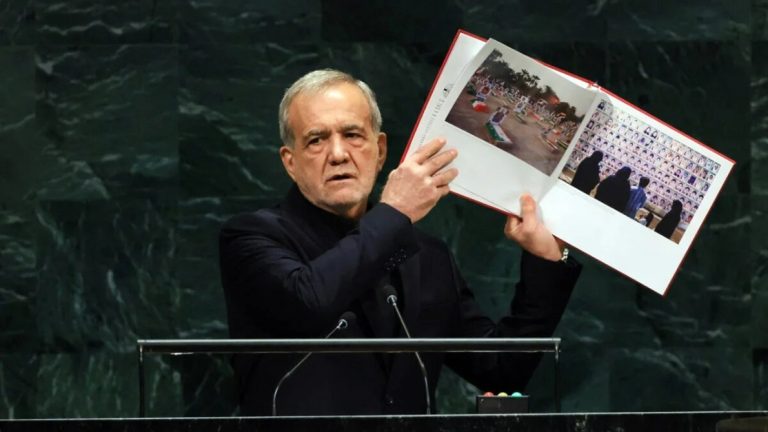 Iranian President Masoud Pezeshkian holding up an open photo book at the UN General Assembly podium.