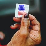 Close-up of a person holding an 'I Voted' sticker with the American flag design between their fingers.
