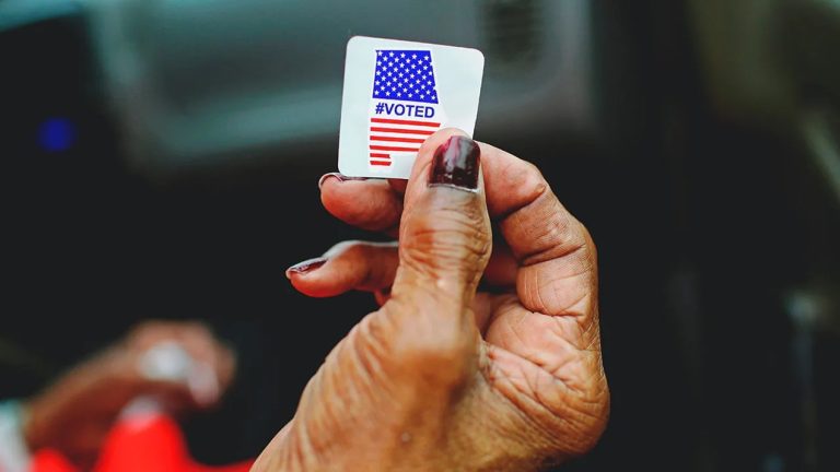 Close-up of a person holding an 'I Voted' sticker with the American flag design between their fingers.
