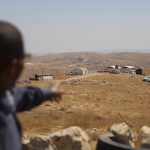 A person pointing toward remote buildings and outposts on a dry, hilly landscape.