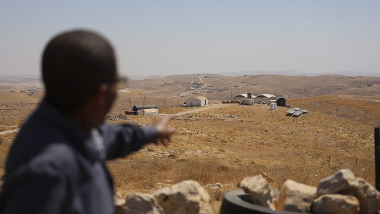 A person pointing toward remote buildings and outposts on a dry, hilly landscape.