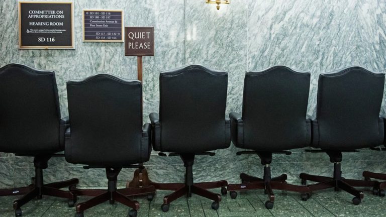 Row of empty black executive chairs outside a Senate committee hearing room.