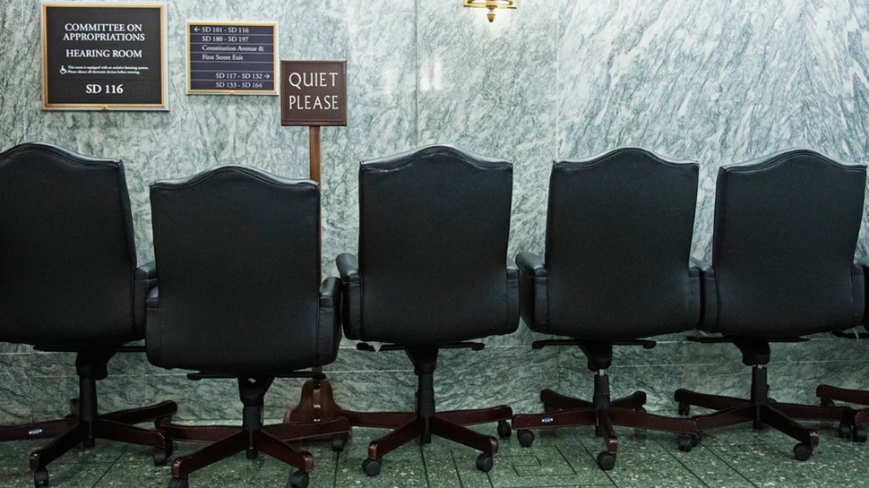 Row of empty black executive chairs outside a Senate committee hearing room.