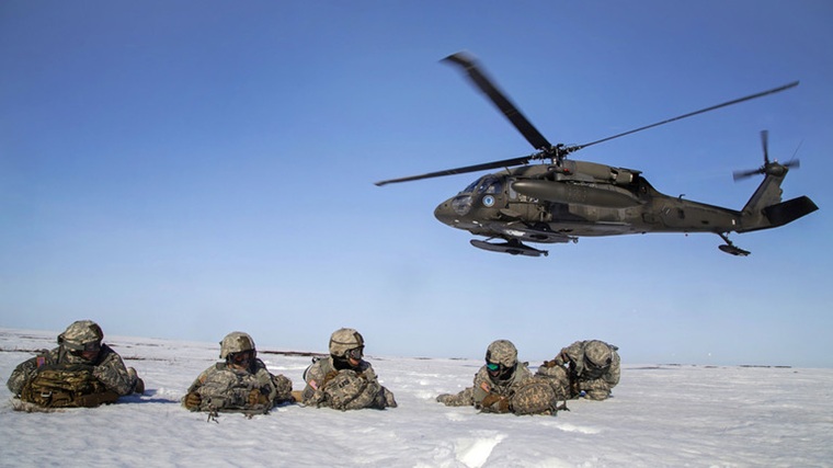 Military helicopter flying low over soldiers lying in the snow.