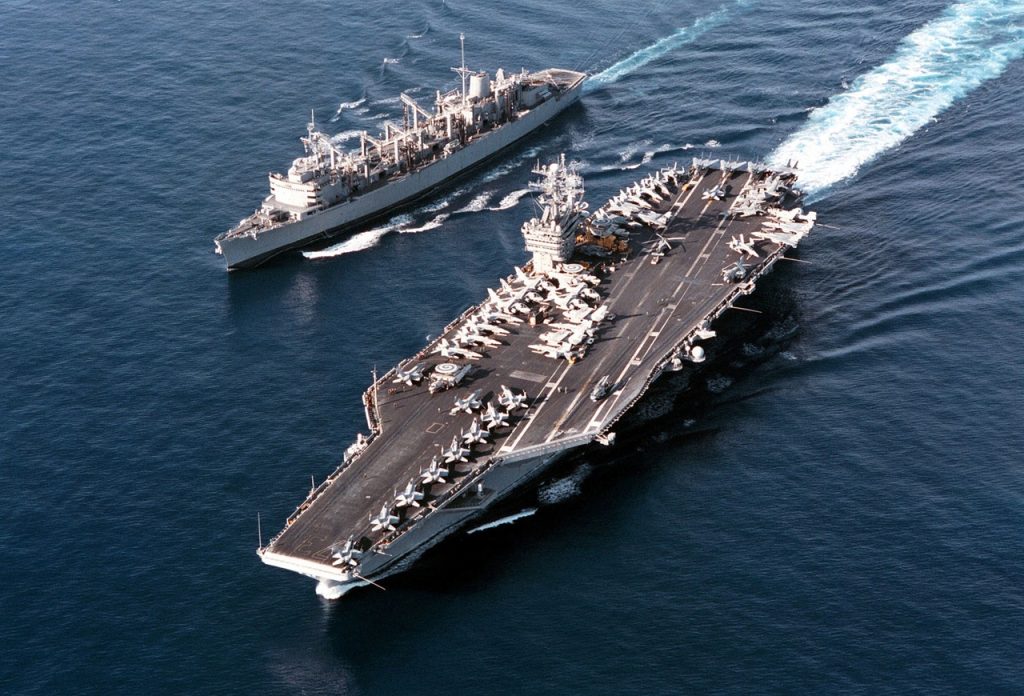 Aerial view of a US Navy aircraft carrier sailing alongside a supply replenishment ship in open ocean.