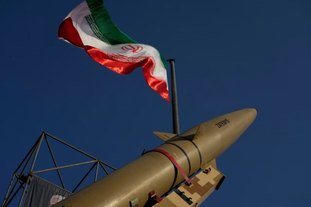 Iranian ballistic missile displayed against a clear blue sky beneath the waving national flag of Iran.