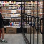 A woman holding a shopping basket looking thoughtfully at egg cartons in a supermarket refrigerated section.