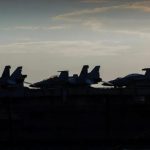 Silhouetted fighter jets lined up on the flight deck of an aircraft carrier at dusk.