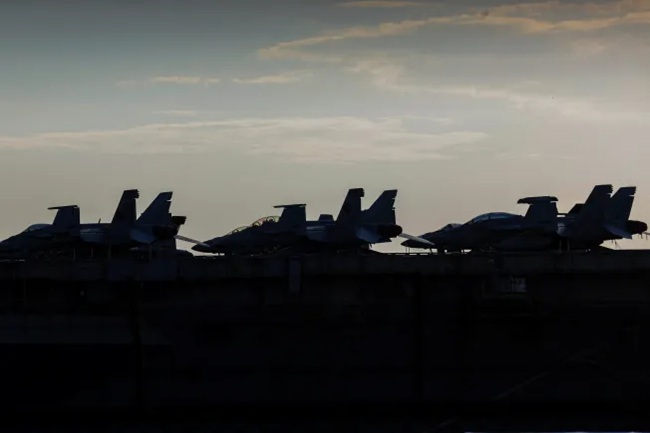 Silhouetted fighter jets lined up on the flight deck of an aircraft carrier at dusk.