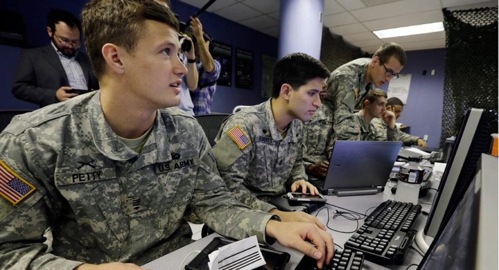 US Army personnel sitting at workstations operating computers in a tech control room.