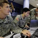 US Army personnel sitting at workstations operating computers in a tech control room.