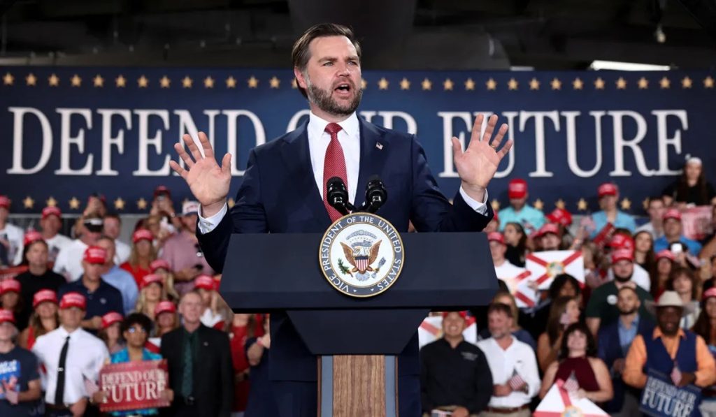 JD Vance speaking at a podium with hands raised in front of a crowd holding campaign signs.