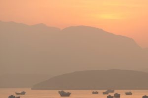 Small boats navigating calm coastal waters at sunset with hazy mountains in the background, representing asymmetric warfare in the Iran conflict.