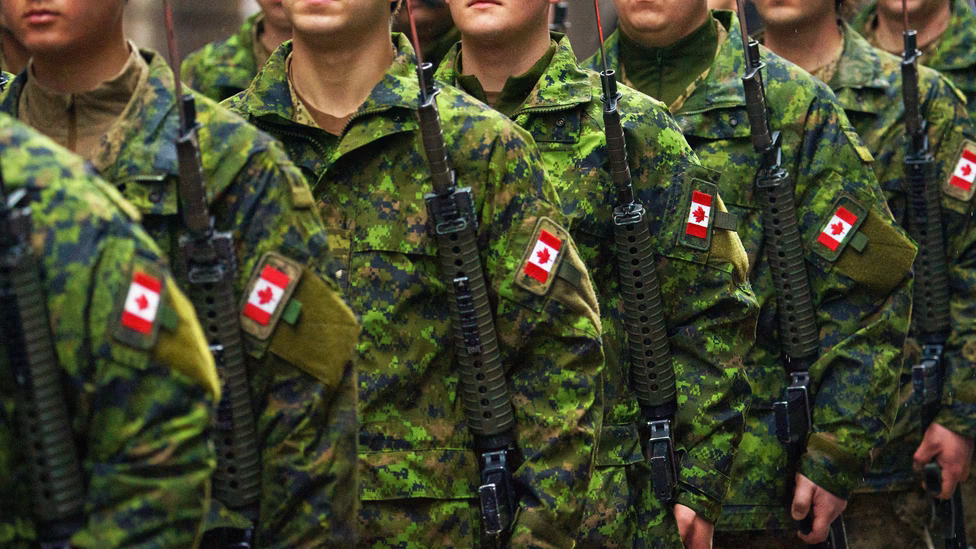 Canadian Armed Forces soldiers in green digital camouflage uniforms standing in formation holding rifles, with Canadian flag patches visible on their sleeves.