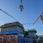 A construction worker in a red hard hat and jacket watches a crane lower a large cylindrical nuclear reactor component at a construction site in China.