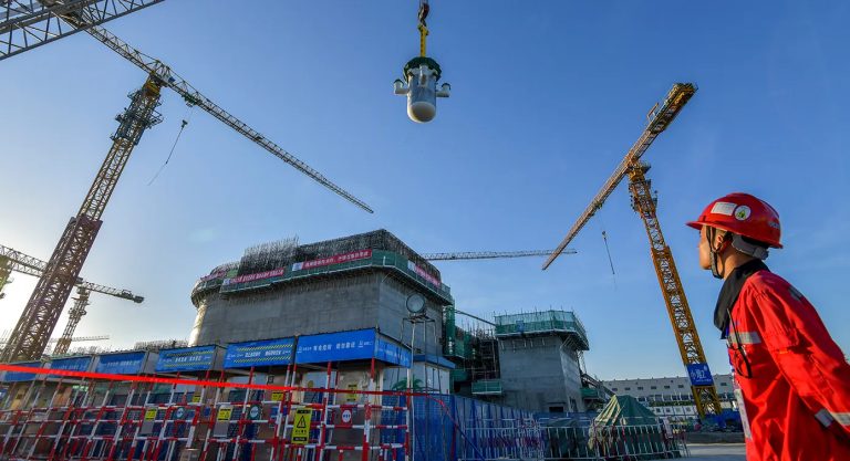 A construction worker in a red hard hat and jacket watches a crane lower a large cylindrical nuclear reactor component at a construction site in China.