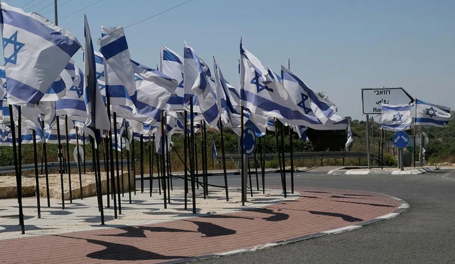Dozens of Israeli national flags fluttering along a roundabout road barrier under a clear sky.
