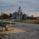 A quiet, rural American landscape featuring an abandoned gas station and old grain silos under a cloudy sky, reflecting economic decline.