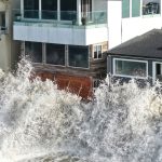 Massive ocean waves crashing violently against the seawall of beachfront homes during a storm.