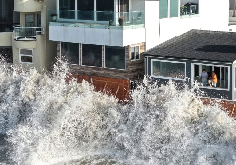 Massive ocean waves crashing violently against the seawall of beachfront homes during a storm.